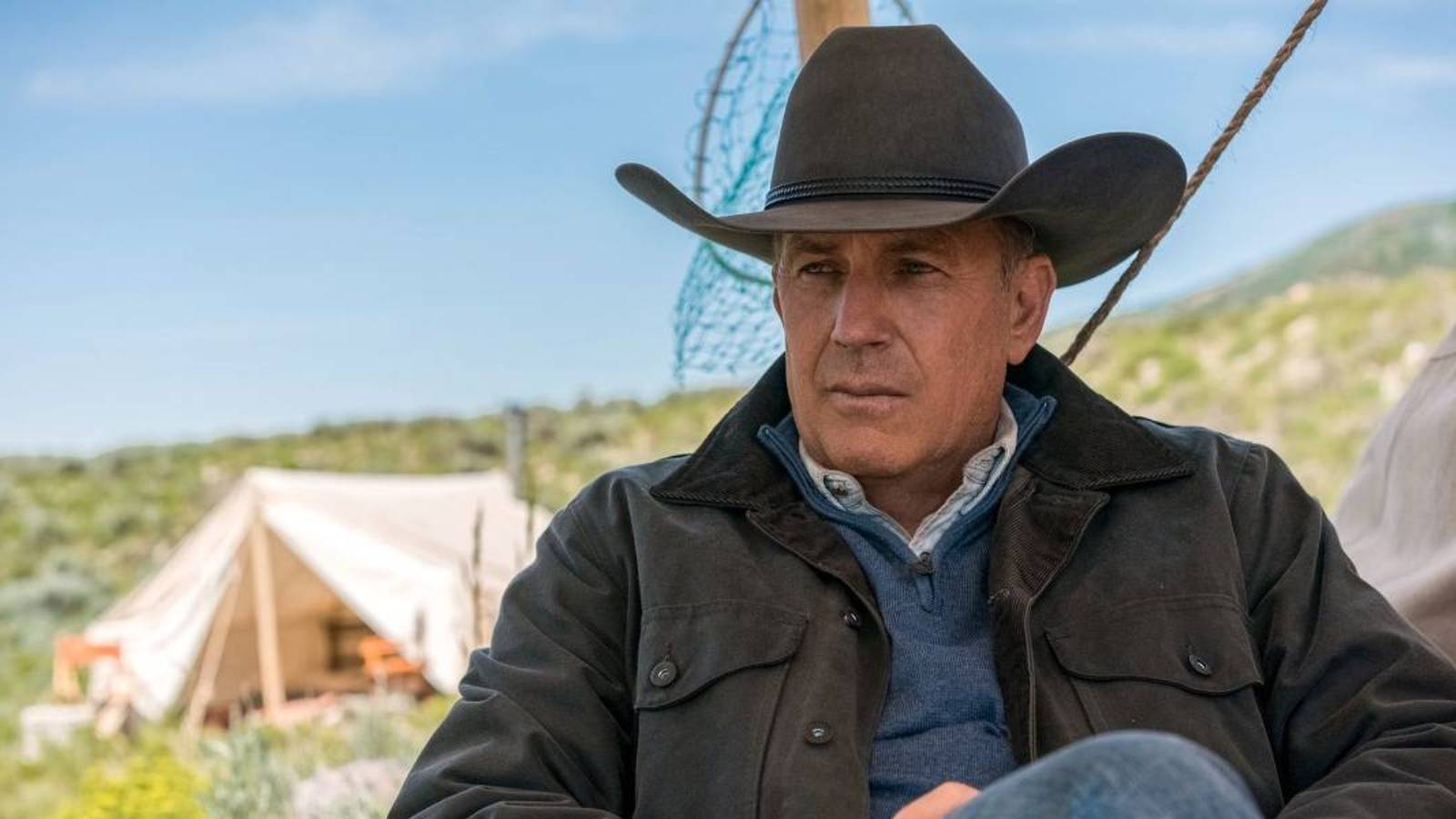 Kevin Costner in Yellowstone sitting with a cowboy hat, a tent in the background.
