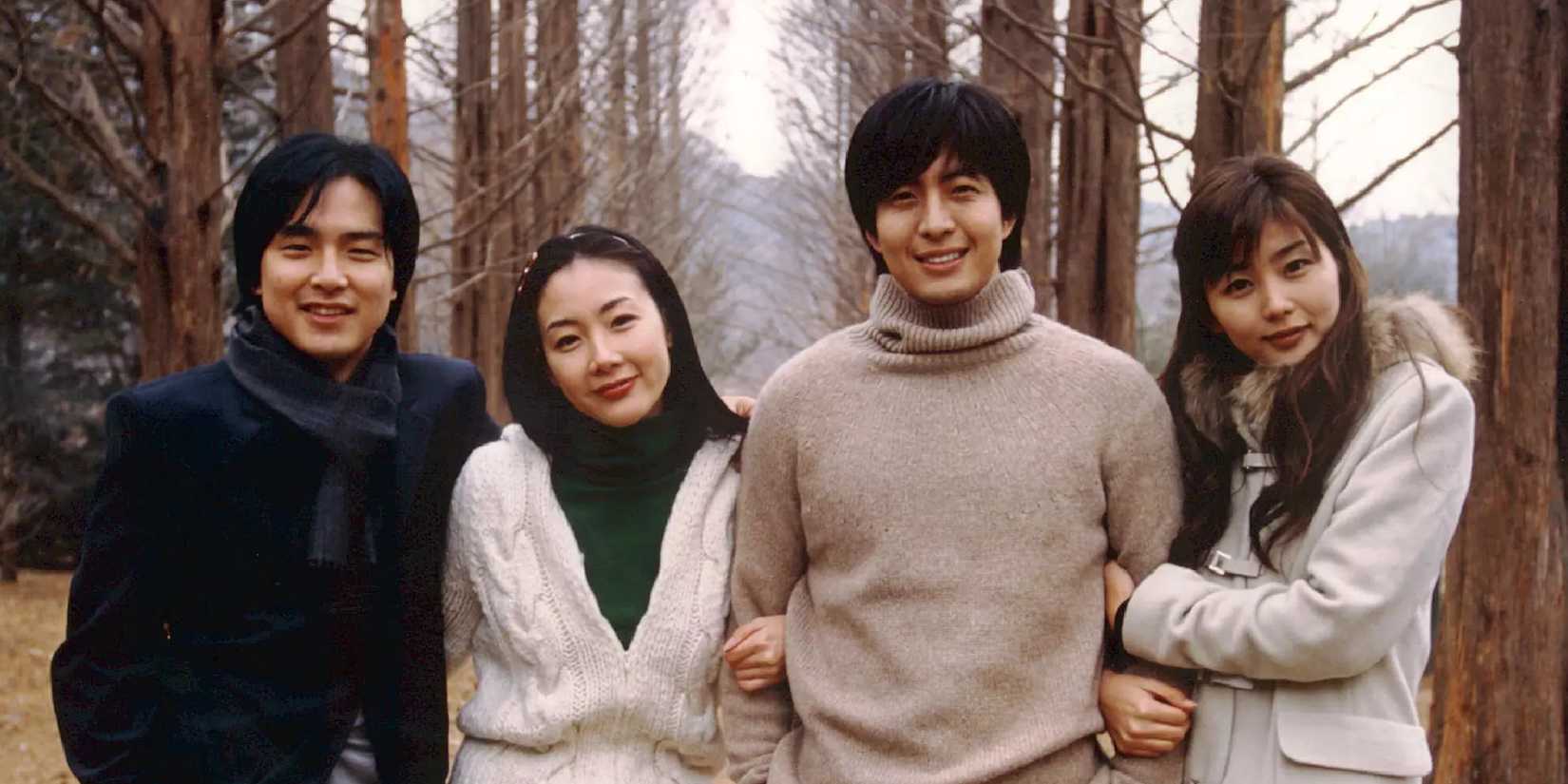 The cast of Winter Sonata wearing winter clothing and smiling at the camera with trees behind.