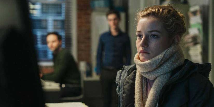 Julia Garner sits at a desk while two men loom behind her and watch her work in The Assistant.
