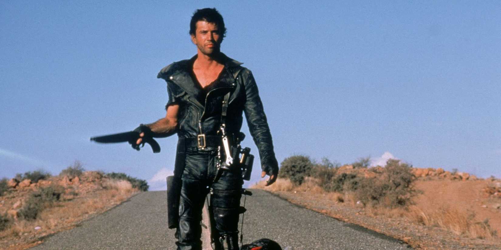 Mel Gibson as Max Rockatansky walking down a lonely highway holding a gun in Mad Max 2 The Road Warrior.