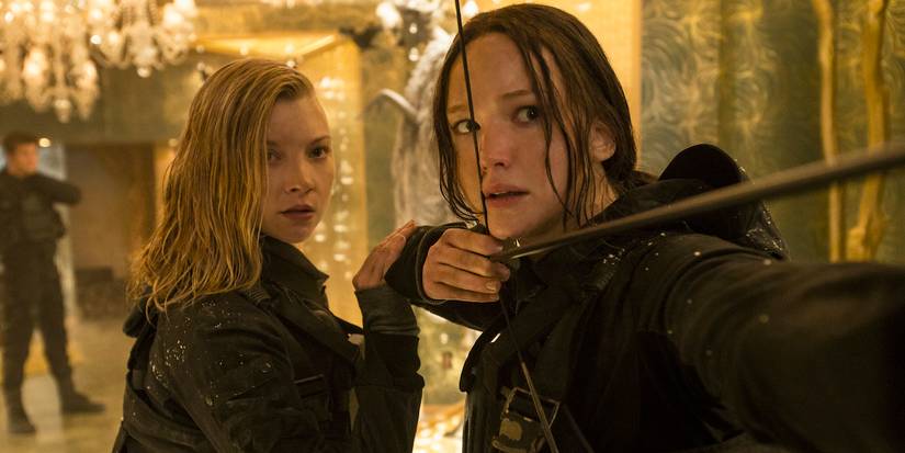 Natalie Dormer next to Jennifer Lawrence drawing a bow and arrow in The Hunger Games: Mockingjay Part 2.