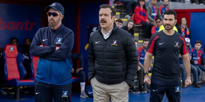 Three of the cast stand near each other and look tense at a sporting event in the Ted Lasso Season 3 finale.