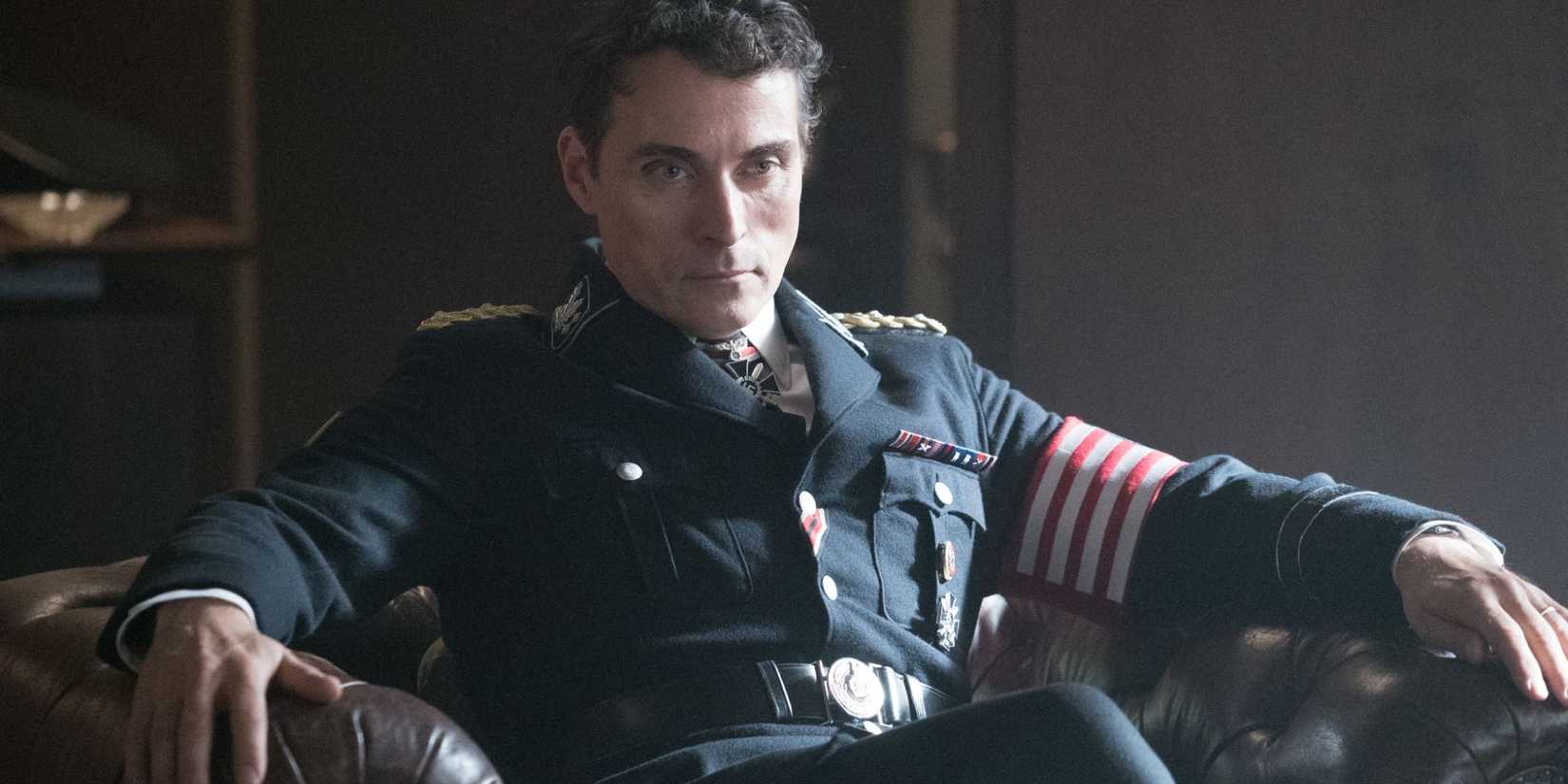 Rufus Sewell as John Smith sits in a chair in a military uniform in The Man in the High Castle
