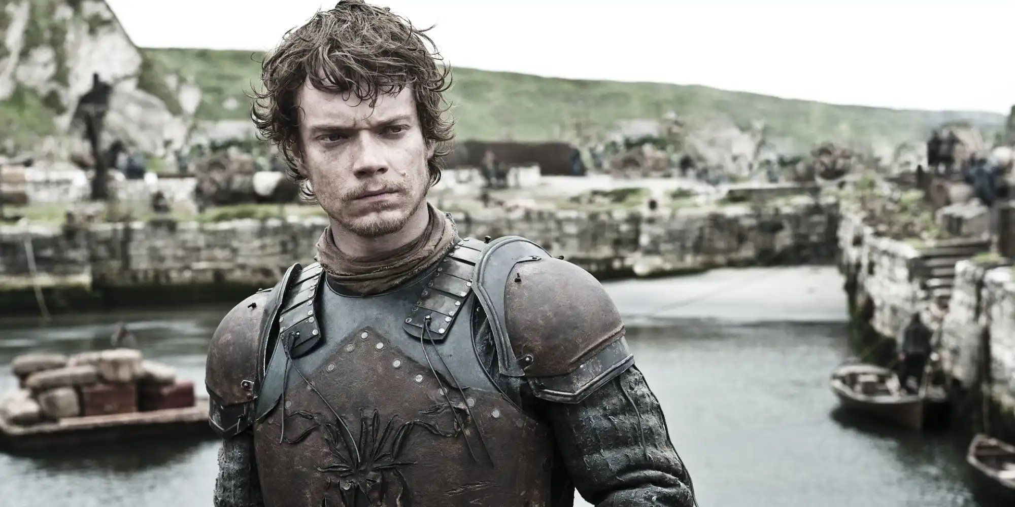 Alfie Allen as Theon Greyjoy standing on a shore looking serious in Game of Thrones.