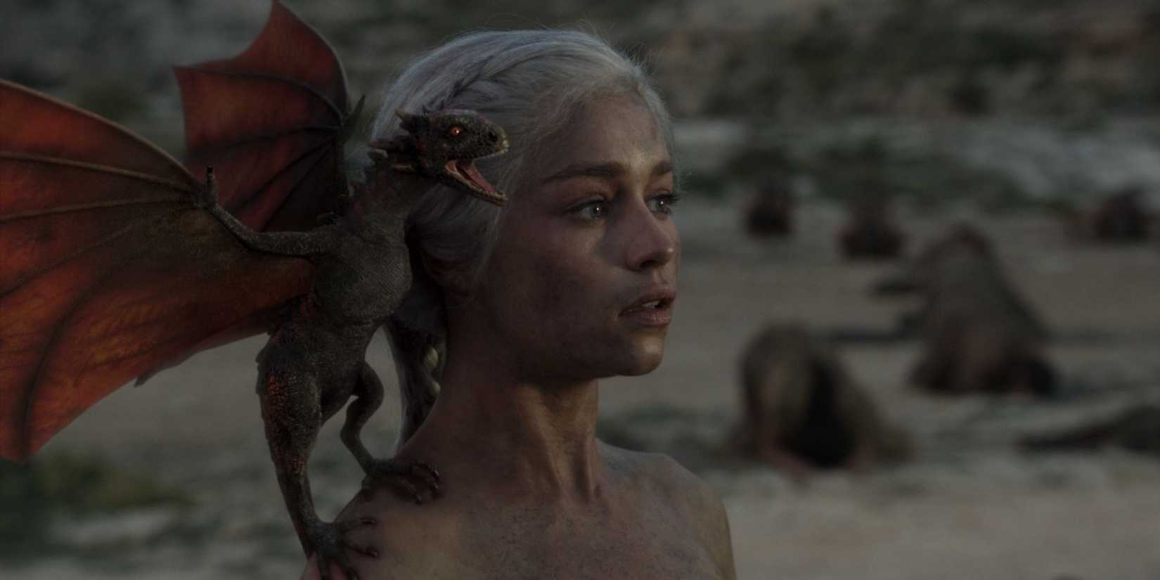 Daenerys Targaryen stands in the ruin of a burnt tent with a newborn baby dragon perched on her shoulder.