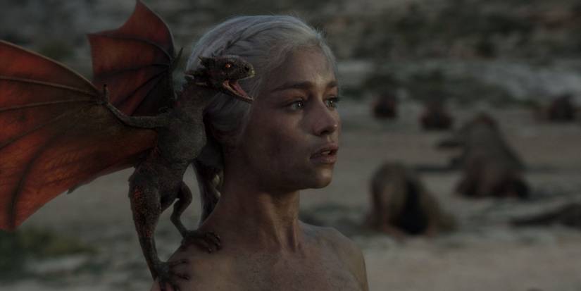 Daenerys Targaryen stands in the ruin of a burnt tent with a newborn baby dragon perched on her shoulder.