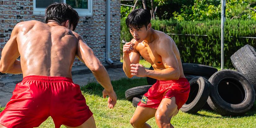 Two men in boxing shorts prepare to fight each other in a yard with grass and old tires in Bloodhounds.