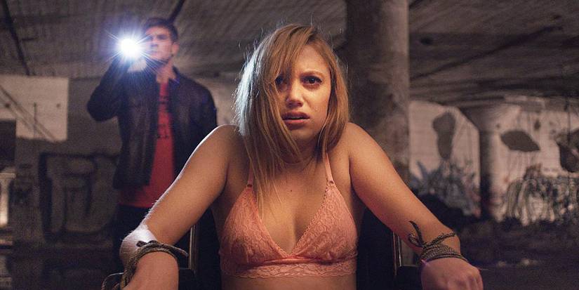 Jay is tied to a chair in her bra with a man with a flashlight behind her in It Follows.