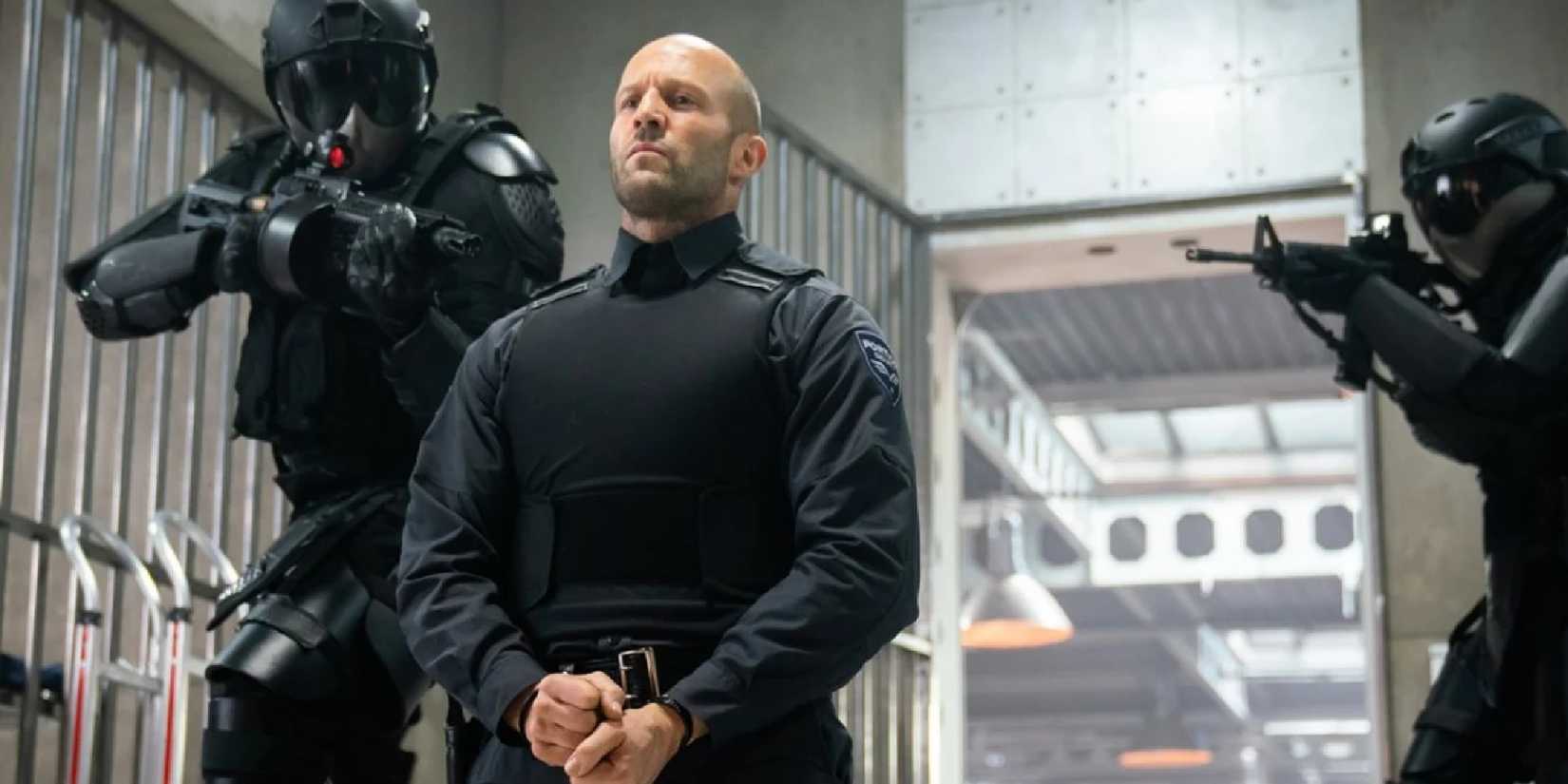 Jason Statham as H with his hands cuffed and soldiers pointing guns at him in Wrath of Man.