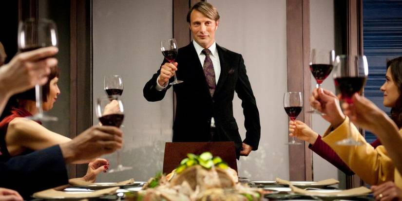 Mads Mikkelsen stands, holding up a glass of wine with others seated around a dinner table in Hannibal.