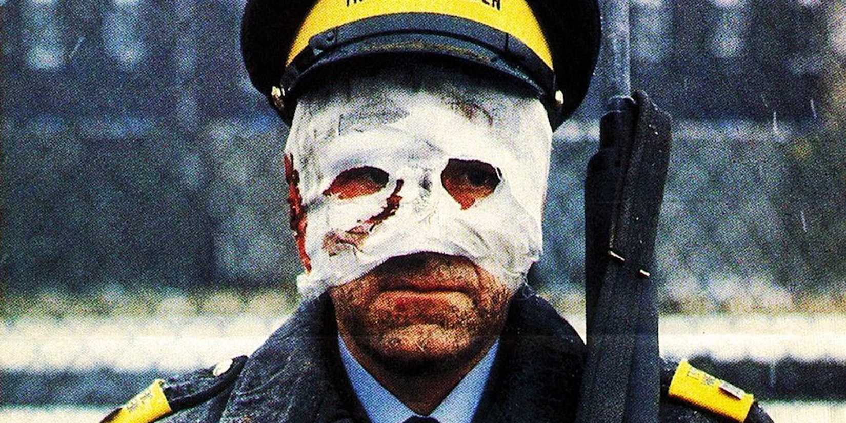 A police officer holds a rifle over his shoulder with a mostly bandaged face in Threads.