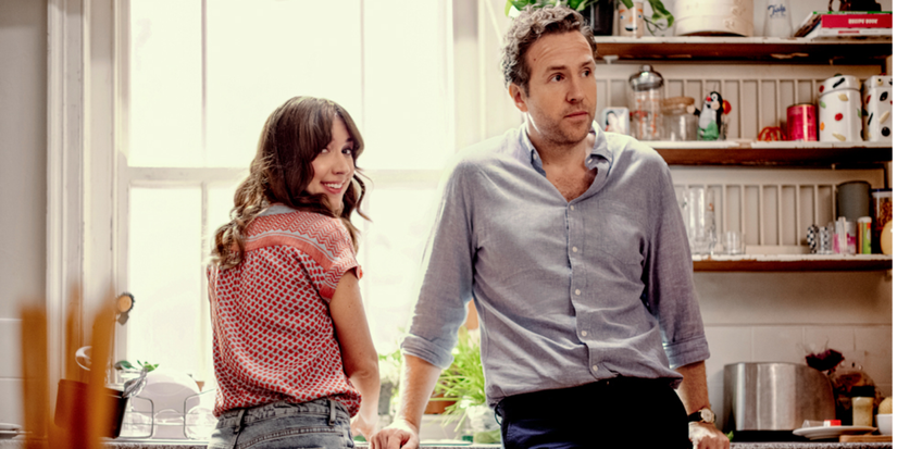 Esther Smith as Nikki and Rafe Spall as Jason standing at a kitchen sink and looking behind in Trying.