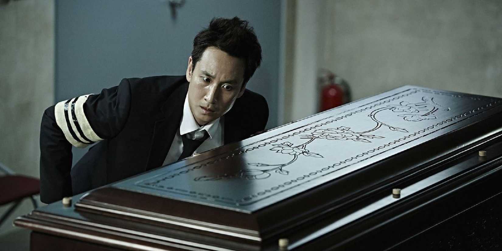 Lee Sun-kyun wearing a suit and a black and white mourning ribbon on his right arm, leaning toward a closed casket with his left ear