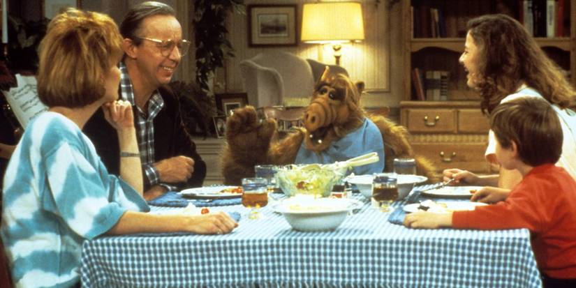 The Tanner family (played by Max Wright, Anne Schedeen, Andrea Elson, and Benji Gregory) sitting at the table with ALF (voiced by Paul Fusco) on 'ALF'