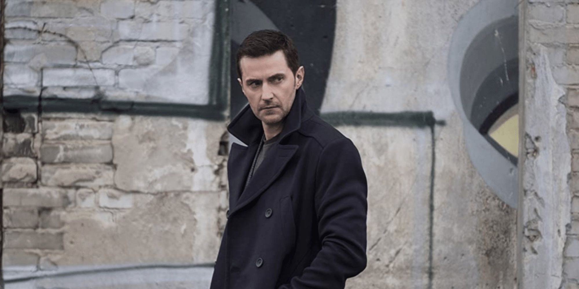 Richard Armitage wears a black trench coat and walks outside looking cautious in Berlin Station.