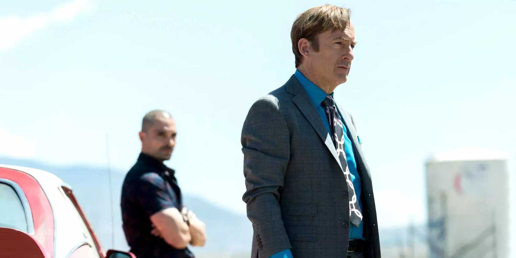 Bob Odenkirk as Saul looking to the distance with a man leaning on a car in the background in Better Call Saul.