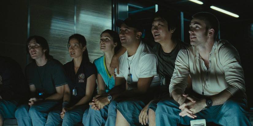 A team of scientists on sit in a rec room on their spaceship, smiling as they look at something together.