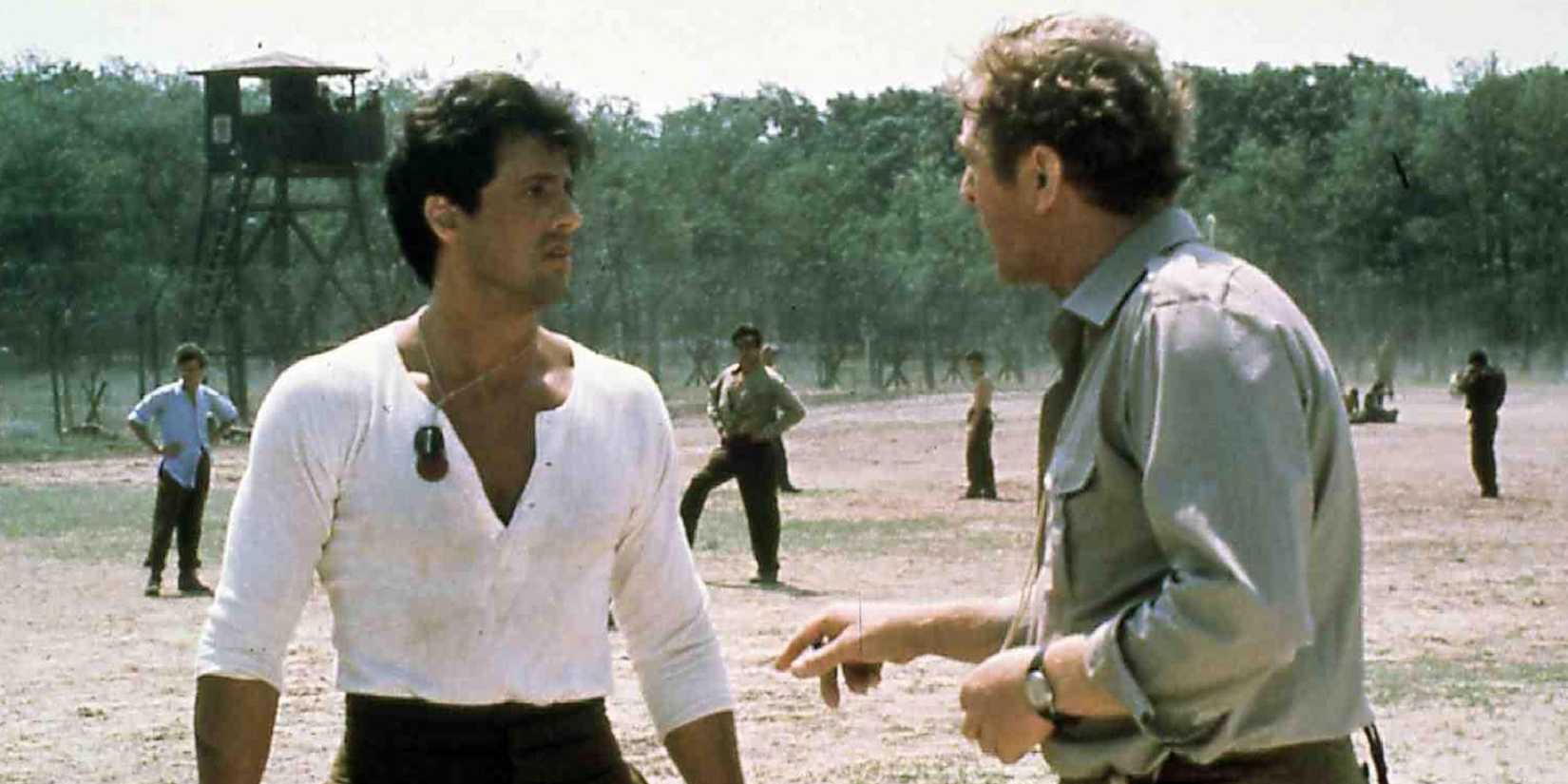 Sylvester Stallone in military dog tags talks to a man with a watchtower behind them in Escape to Victory.