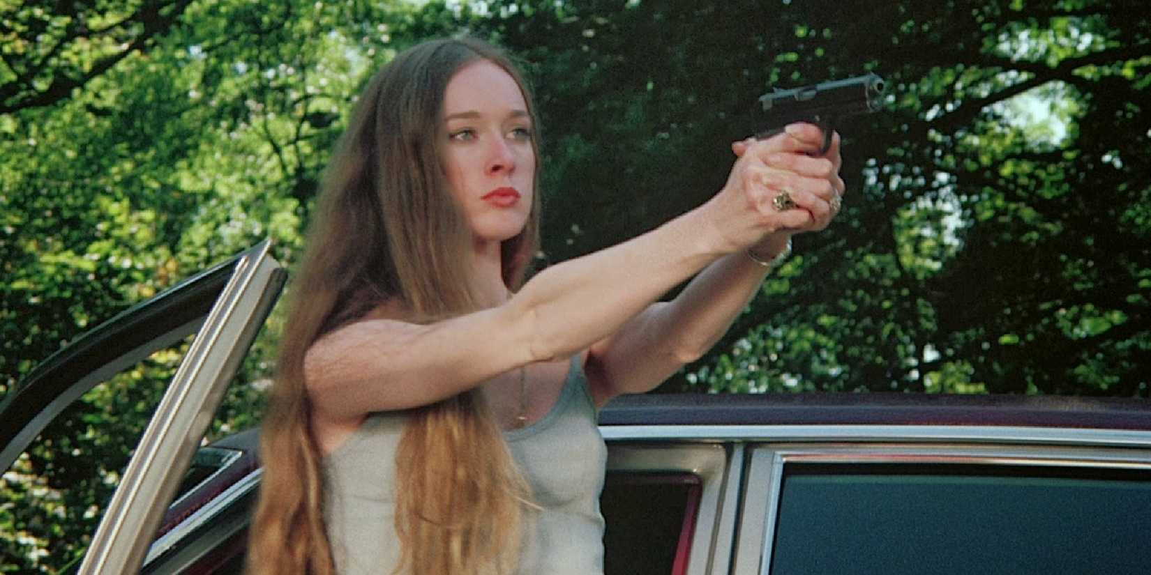 A young woman stands outside her car door pointing a gun at someone off-camera