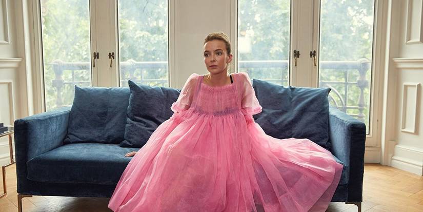 Jodie Comer sebagai Villanelle dalam gaun berwarna merah muda permen karet duduk di sofa menatap ke angkasa di Killing Eve.