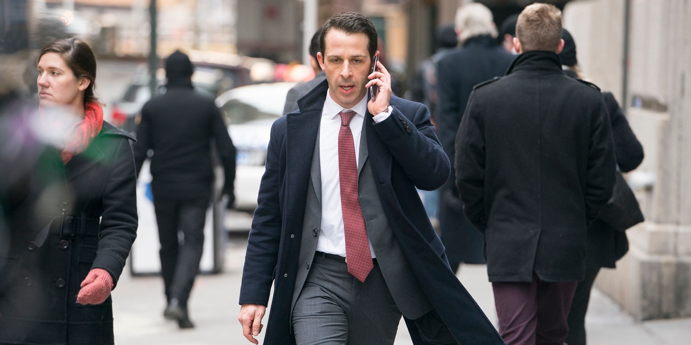 Kendall Roy from Succession walking down the street in a peacock and suit on the phone looking angry.
