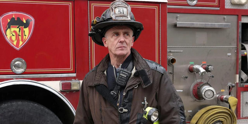 David Eigenberg wearing firefighter gear, standing in front of a fire truck in Chicago Fire.