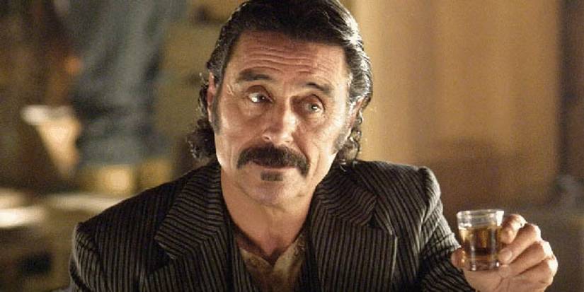 Ian McShane as Al Swearengen holding up a shot of liquor in Deadwood. 