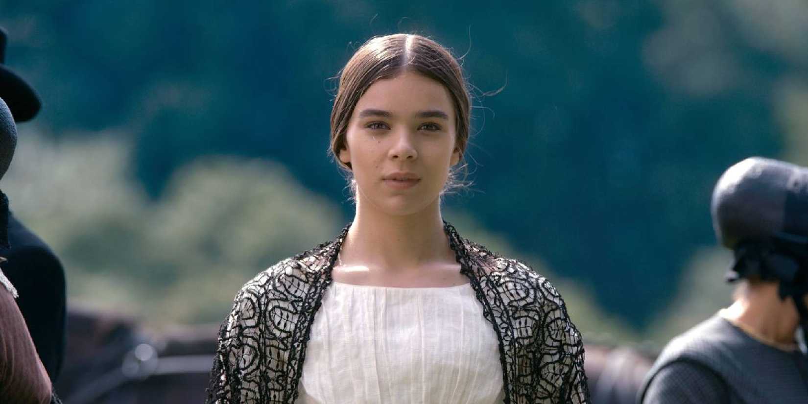 Hailee Steinfeld wears a period costume and stands outside with people going past her in Dickinson.
