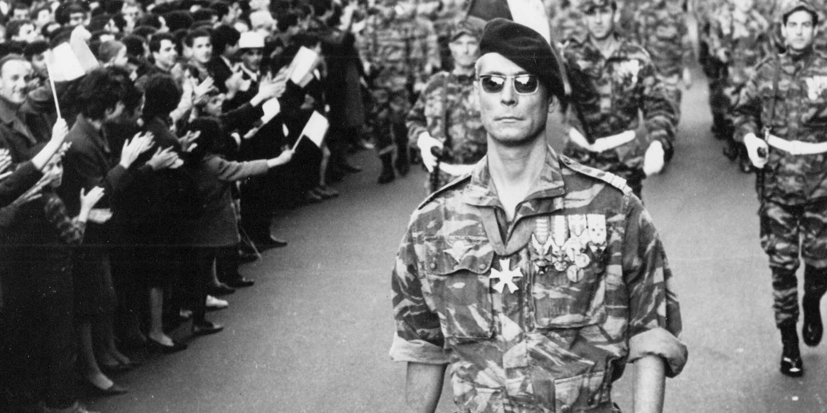 A soldier walking in front of several other soldiers while bystanders look in The Battle of Algiers