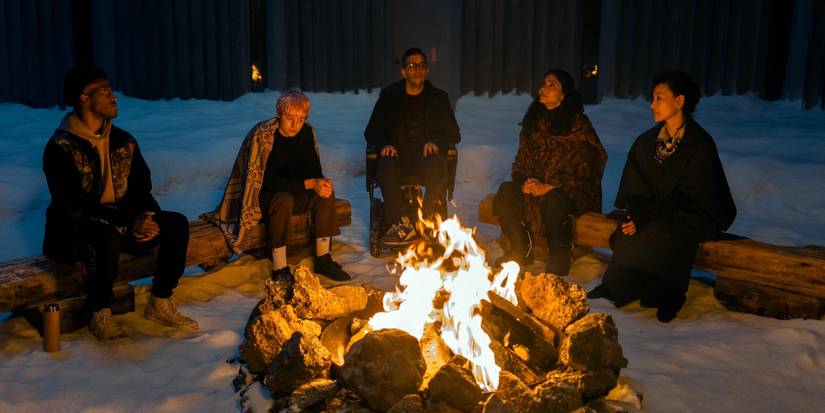 People sit on log benches around a fire in the snow in A Murder at the End of the World. 