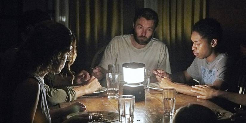 three people sitting at a table around a lamp in It Comes at Night 