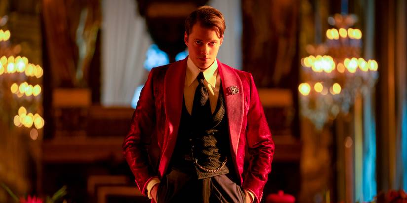 Bill Skarsgård as The Marquis de Gramont stands in a decadent room in a lavish red suit.