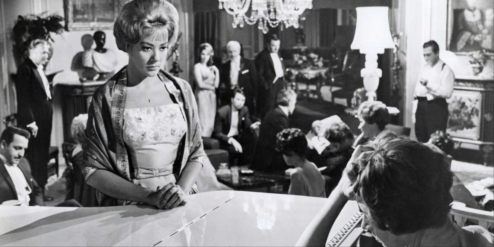 Silvia Pinal as Leticia 'La Valkiria' standing by a piano with a crowd of people behind her in The Exterminating Angel