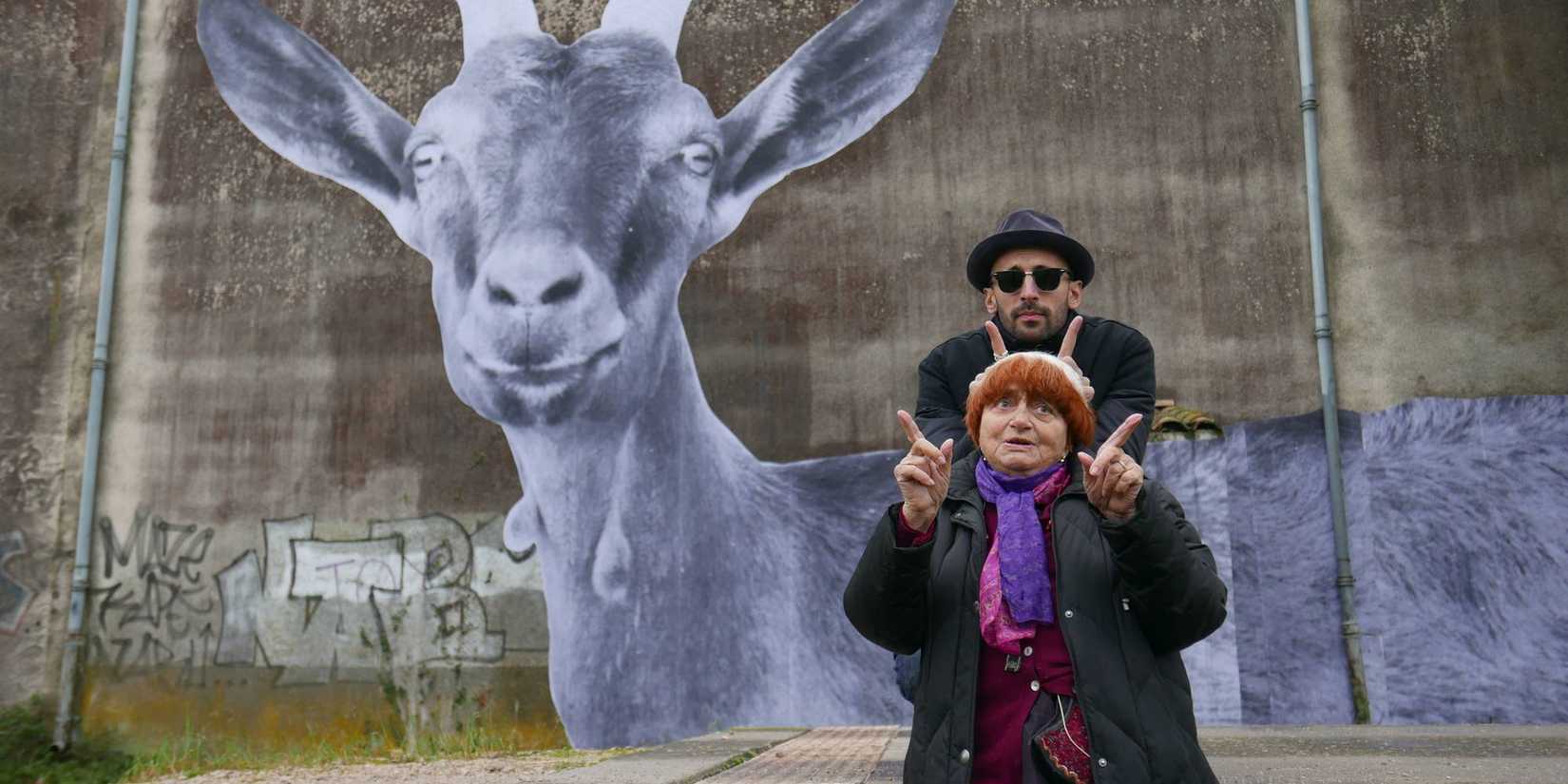 JR and Agnès Varda making silly faces while posing in front of a wall with a goat print in it in Faces Places