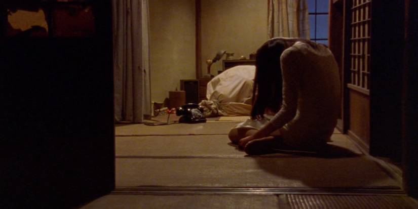 A girl sits hunched on the ground in a room with a phone and a bag on the ground in front of her.