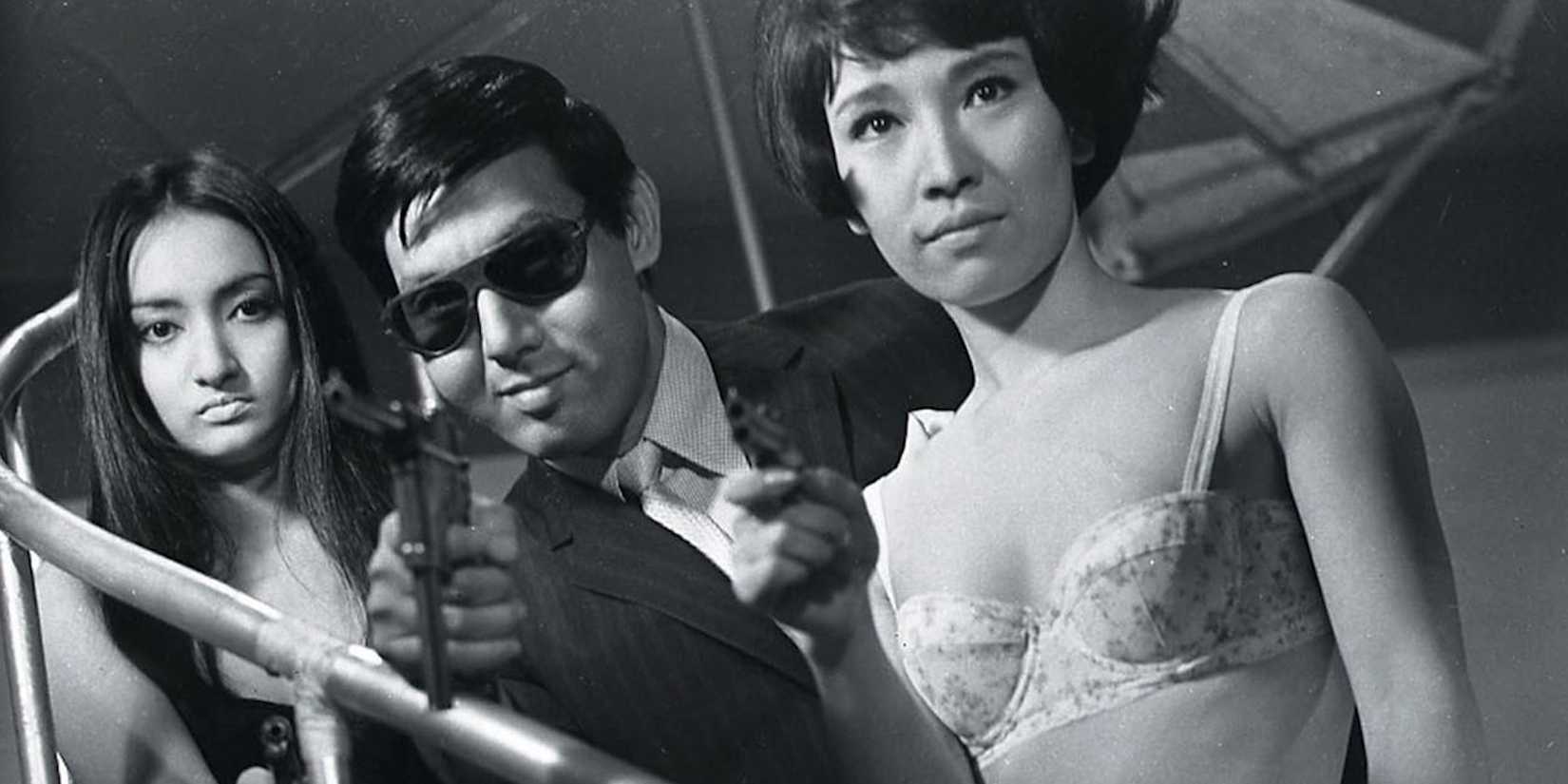 A man aiming a gun to the camera while two women stand besides him in Branded to Kill 1967