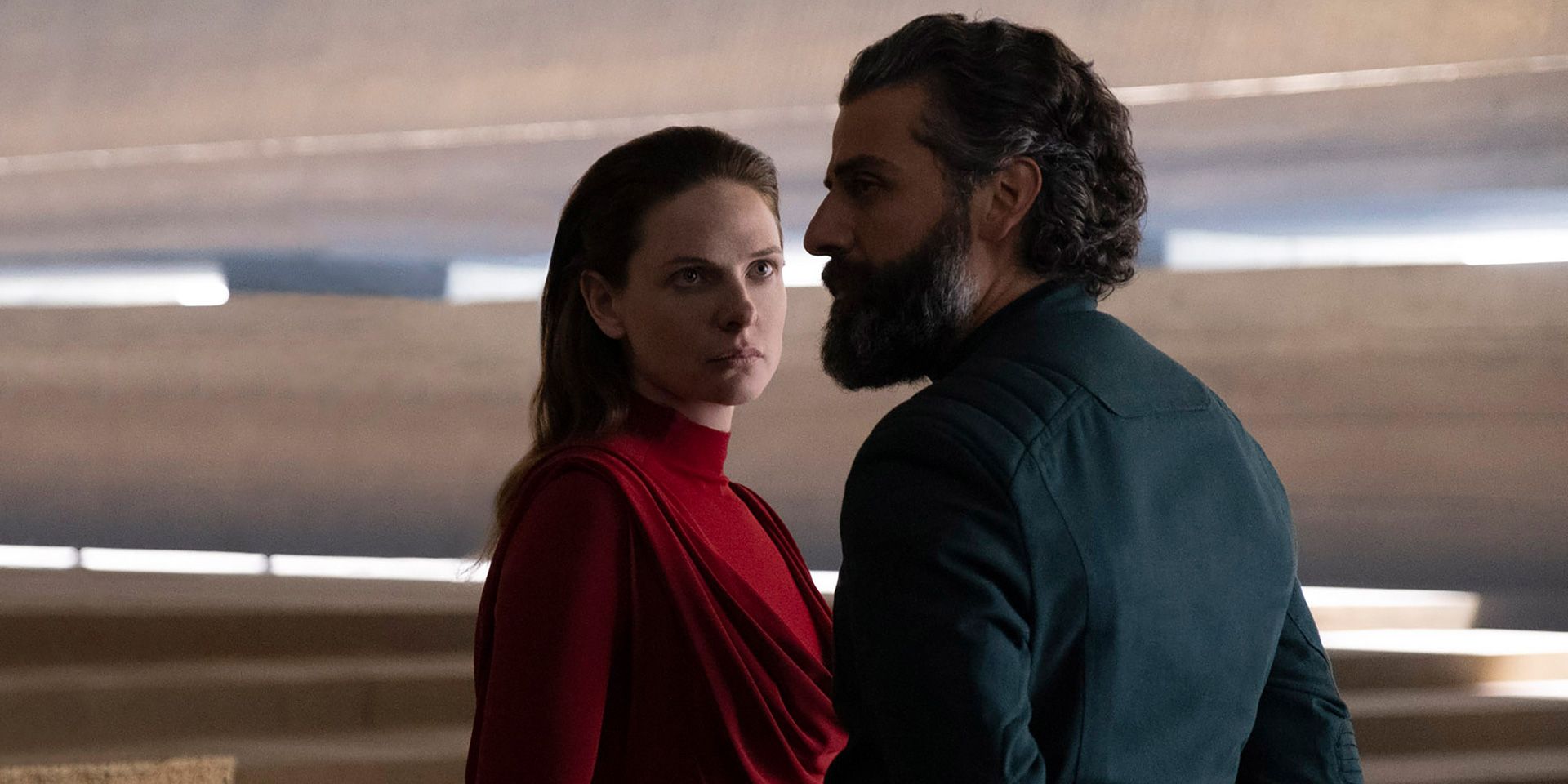 Rebecca Ferguson as Lady Jessica, wearing a red dress with her hair down and looking at Oscar Isaac's Duke Leto Atreides, wearing a dark suit and looking away from her to the left, in Dune