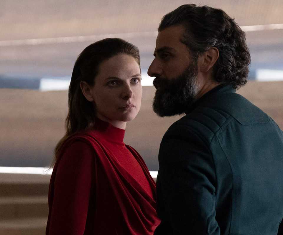 Rebecca Ferguson as Lady Jessica, wearing a red dress with her hair down and looking at Oscar Isaac's Duke Leto Atreides, wearing a dark suit and looking away from her to the left, in Dune