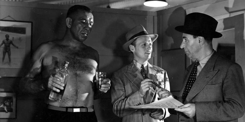 A con artist hands a pen and paper to someone while a large man stand shirtless beside him pouring a drink.