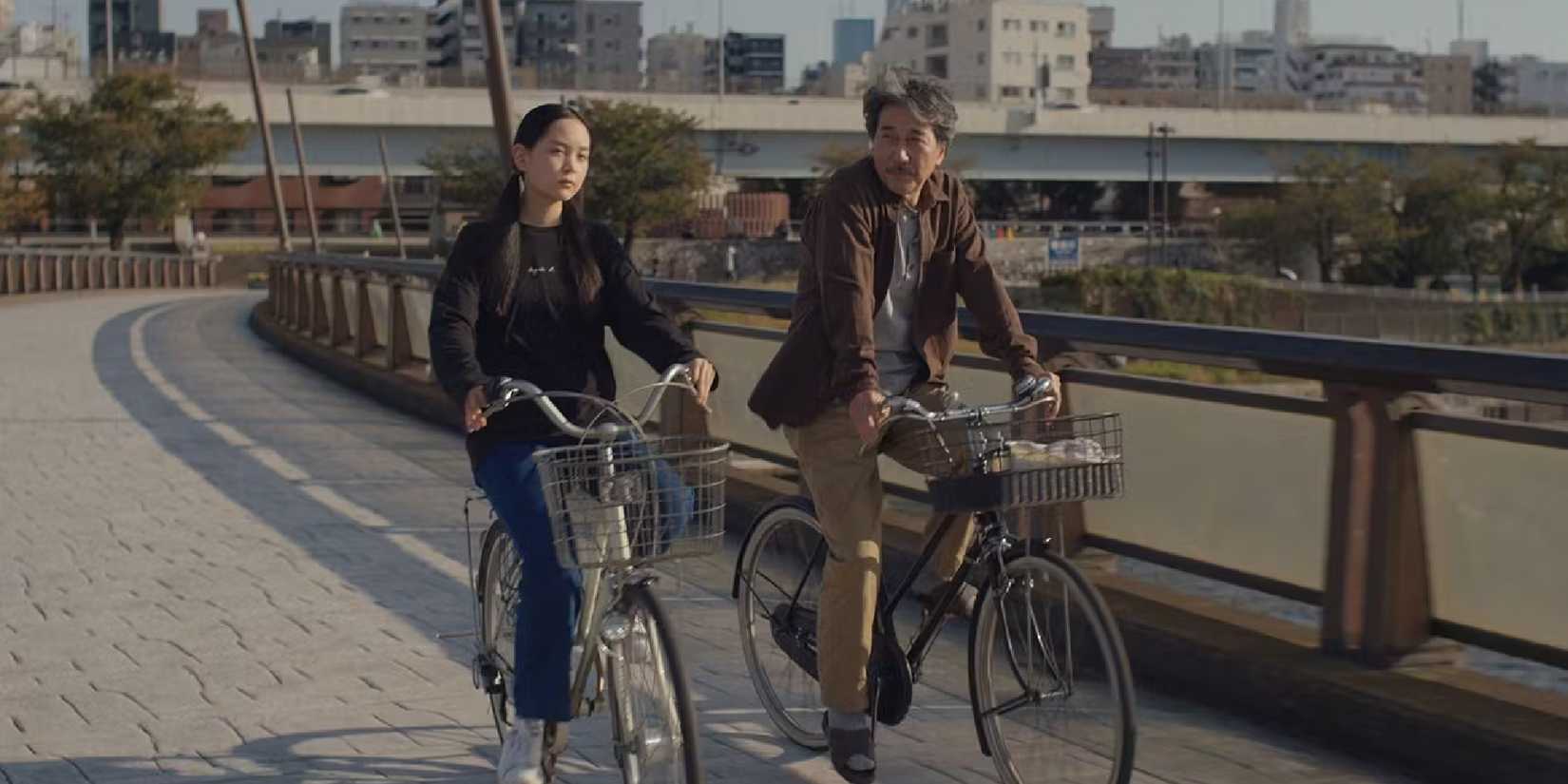 A man and woman ride bicycles on a street in a city in Perfect Days.
