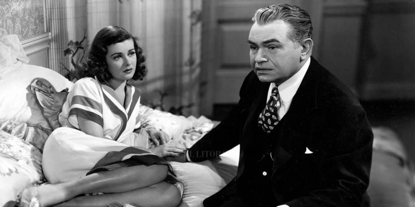 A man and a woman sit on a bed holding hands with resigned looks on their faces.