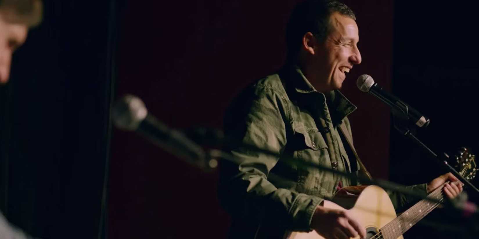 Adam Sandler smiling on stage with a guitar for his stand-up special 100% Fresh on Netflix