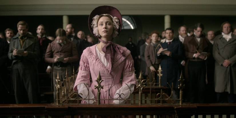 Sarah Gadon as Grace Marks standing up in the courtroom looking straight ahead and wearing a pink dress and bonnet, with a crowd standing behind her, in Alias Grace