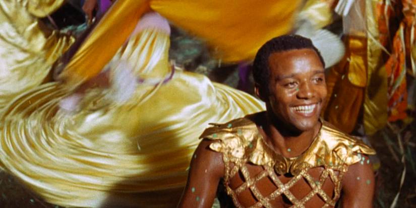 Orfeu (Breno Mello) smiles gleefully as a woman in a vibrant yellow dress twirls behind him. 