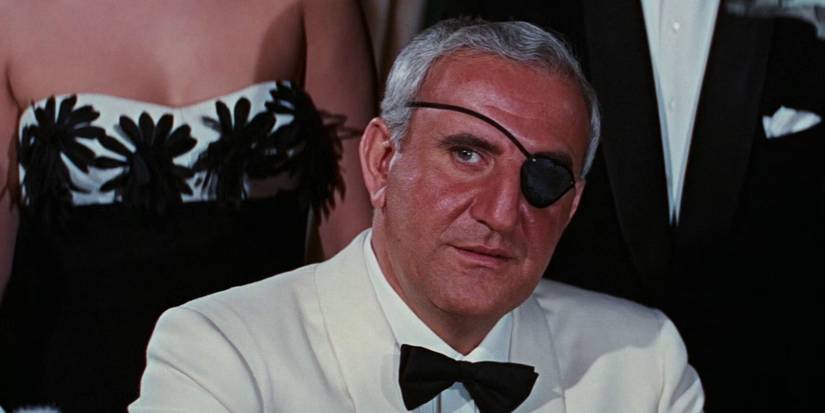 Bond villain Emilio Largo sits at a table in a white tuxedo as people stand behind him in Thunderball