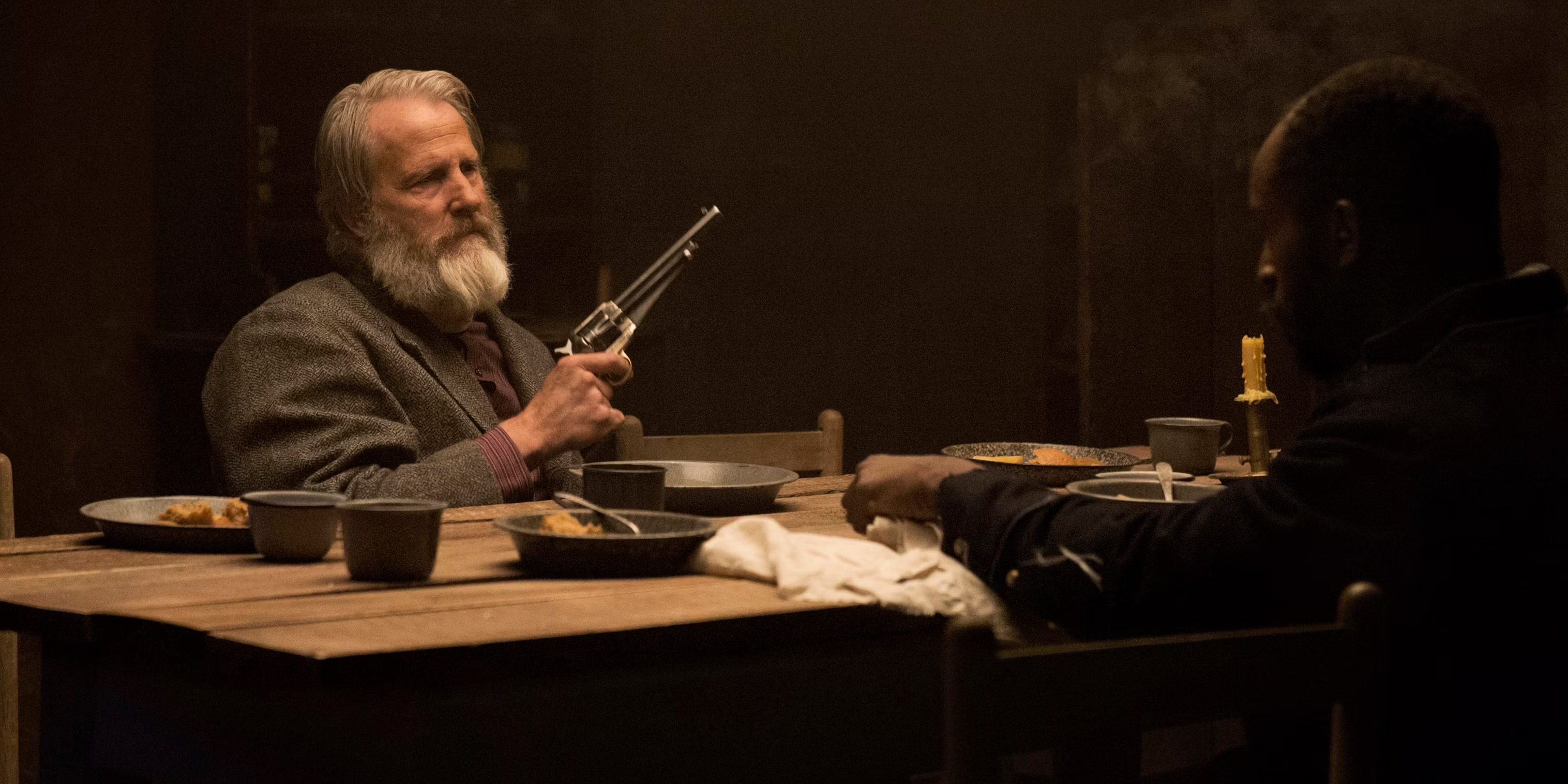 Frank Griffin (Jeff Daniels) holding a gun up while sitting at a table in Godless