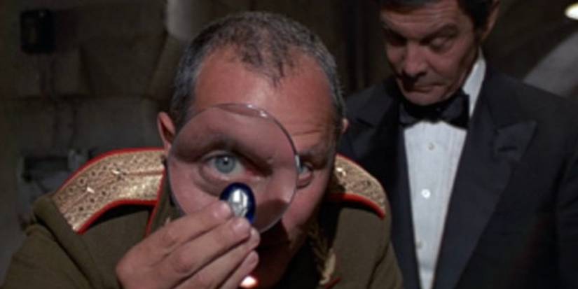 A Soviet military man looks at a diamond through a magnifying glass while a crooked smuggler stands behind him in a tuxedo.