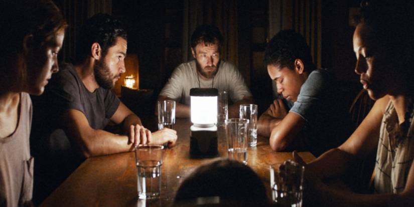 The survivors gather around a table with a single lamp in the middle in It Comes at Night.