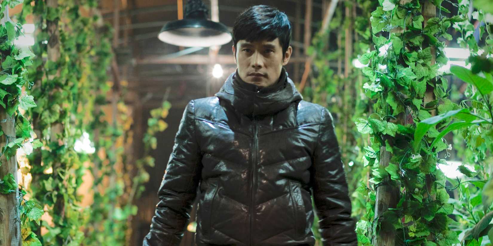 Lee Byung-hun sebagai Kim Soo-hyun tampak serius, berdiri di antara tanaman budidaya di I Saw the Devil.