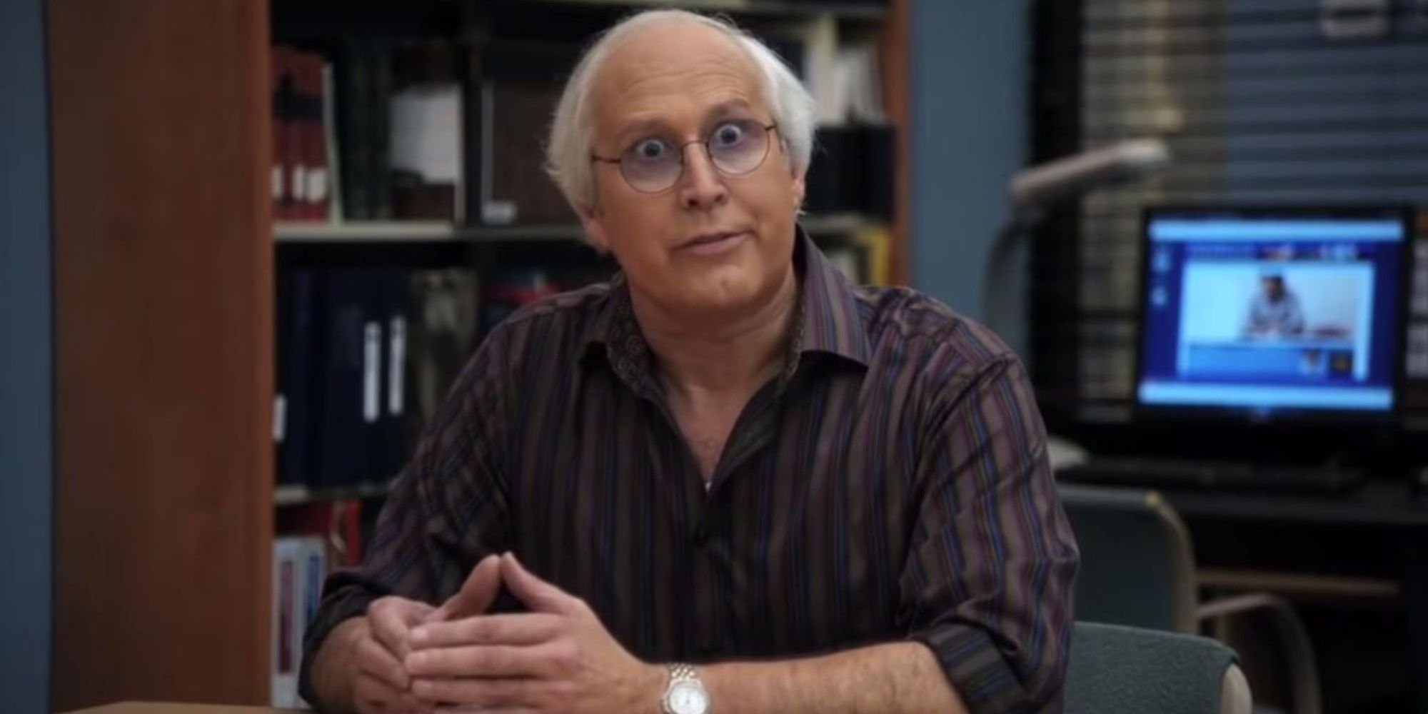 Pierce Hawthorne (Chevy Chase) de Community está sentado con las manos cruzadas en una mesa de su laboratorio.